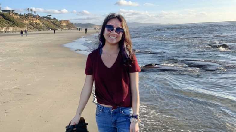 Student standing on a beach, near the water.