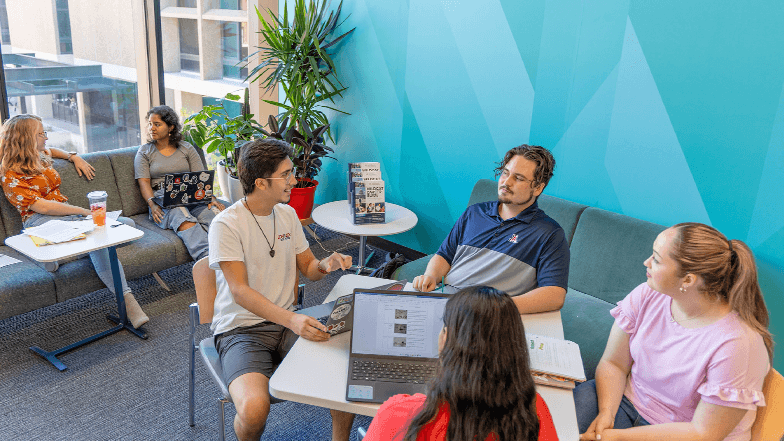 A group of University of Arizona students is gathered around a table talking in an on-campus room. 