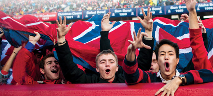 Students cheering in the ZonaZoo section under a wildcat flag at a football game.