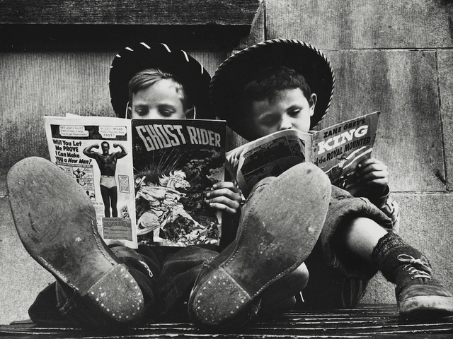 Two boys in cowboy hats sit side by side with their feet stretched out toward the camera, each reading a comic book. One holds The Ghost Rider while the other reads a Zane Grey western. The image is in black and white, highlighting their worn shoes and absorbed expressions.