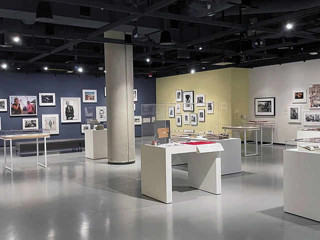 A modern gallery space at the Center for Creative Photography featuring an exhibition of framed photographs and archival materials. The walls are painted in dark blue, soft yellow, and white, with artworks arranged in salon-style groupings. Display cases throughout the room contain books, prints, and other photographic items under glass.