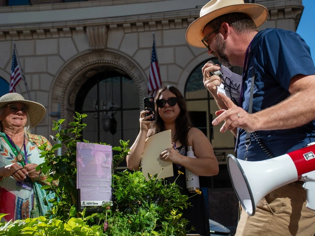 A man in a blue shirt and sun hat speaks into a megaphone while reading a poem printed on a plastic plaque, placed in a plant pot. Behind him, a woman with long brown hair films on a cell phone as another woman watches while smiling.