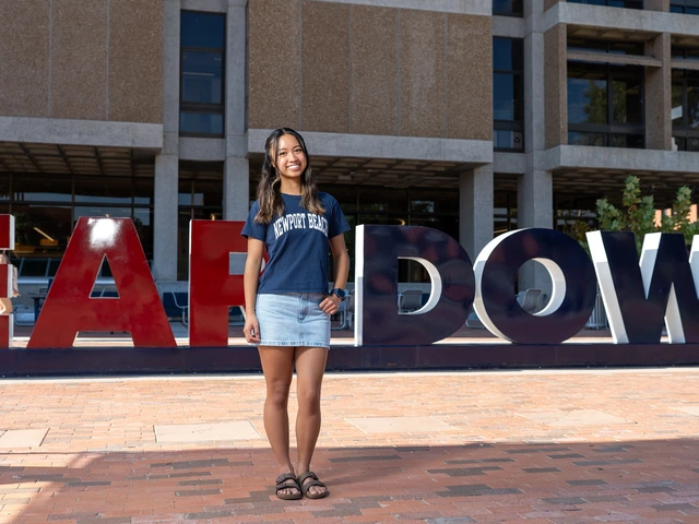 A young woman wearing a navy blue tshirt and denim skirt with long brown hair stands on red brick with one hand on her hip.