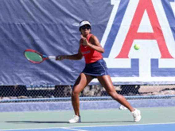 A female tennis player wearing a red tank top, navy athletic shorts, white sneakers, and sunglasses prepares to hit a forehand shot on an outdoor tennis court. The U of A large “A” logo is visible on the blue banner in the background.