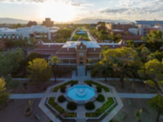 An aerial view of the University of Arizona campus at sunrise, centered on the historic red-brick Old Main building surrounded by green trees and a circular fountain in the foreground. The sun rises over distant mountains, casting a warm glow across the scene.