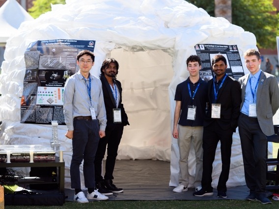 A group of college-aged men wearing semi-formal clothing and wearing name badges pose in front of a white, circular tent on the side of which are informational posters.
