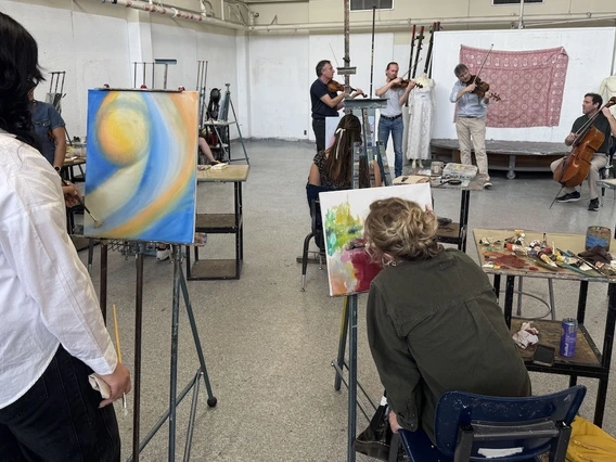 University of Arizona art students paint at easels in a studio while a string quartet performs live music in the background. The students are focused on their colorful abstract paintings, with palettes and paint supplies on nearby tables. The musicians, standing in front of a red tapestry and dress forms, include three violinists and a cellist.