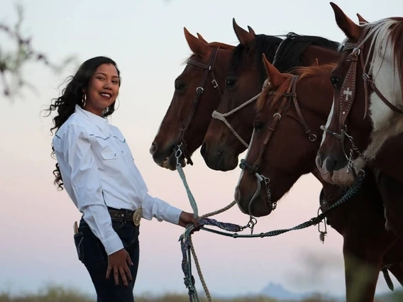 A woman with long dark hair wearing dark denim jeans and a long sleeved white formal shirt stands in front of four horses in an outdoor environment. The sun is setting in the background. The woman is holding leads to all four horses.