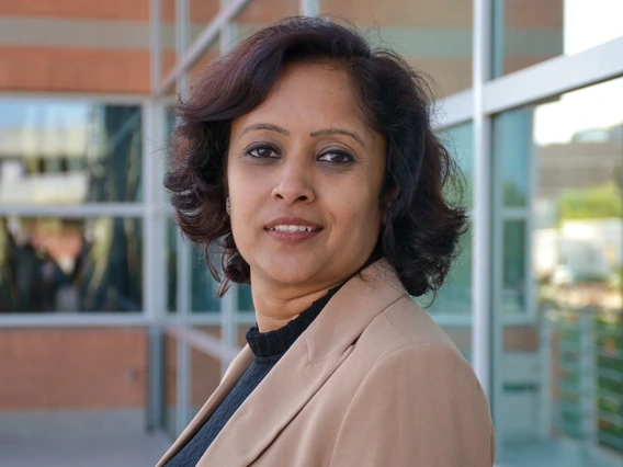 A confident woman with short, wavy dark hair stands in front of a modern glass and brick building. She wears a beige blazer over a black top and gazes calmly into the camera, with natural daylight highlighting her professional presence.