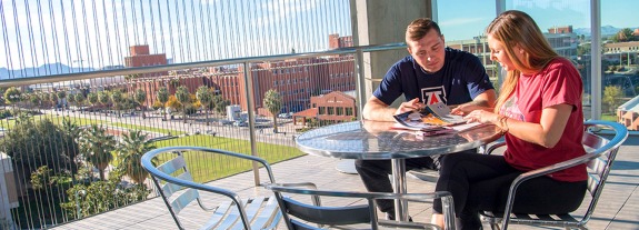 Two Arizona students sitting on chairs reading with a view of campus in background