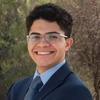 A young man with short curly hair and glasses smiles confidently in a professional outdoor portrait. He wears a navy blue suit, a light blue dress shirt, and a patterned tie, standing against a natural desert background with green and brown foliage.
